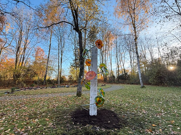 Weiße, schlanke Stele mit bunten,  Glas-Blättern in einer herbstlichen Parklandschaft mit Bäumen und Laub auf dem Boden.