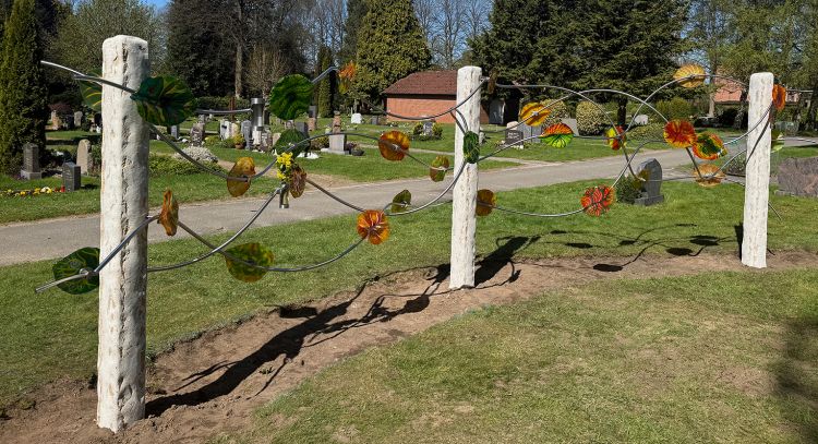 Urnen-Gemeinschafts-Grabanlage mit 30 bunten Glasblättern auf einer grünen Wiese. Im Hintergrund sind Grabsteine zu sehen. Urnen-Gemeinschafts-Grabanlage mit 30 bunten Glasblättern auf einer grünen Wiese. Im Hintergrund sind Grabsteine zu sehen.