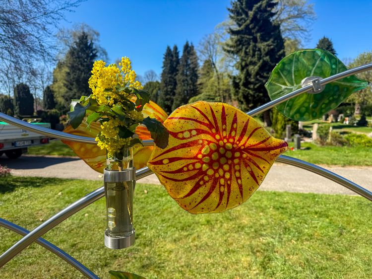 Buntes Glasblatt, dass von der Sonne hell erleuchtet wird. Im Hintergrund sind Grabsteine zu sehen.