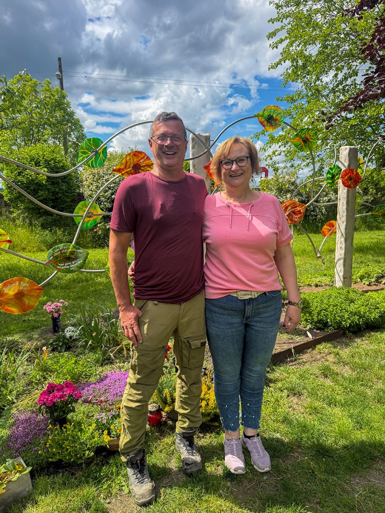 Eine Frau und ein Mann stehen vor einer Urnen-Gemeinschafts-Grabanlage mit bunten Glasblättern und strahlen in die Kamera.