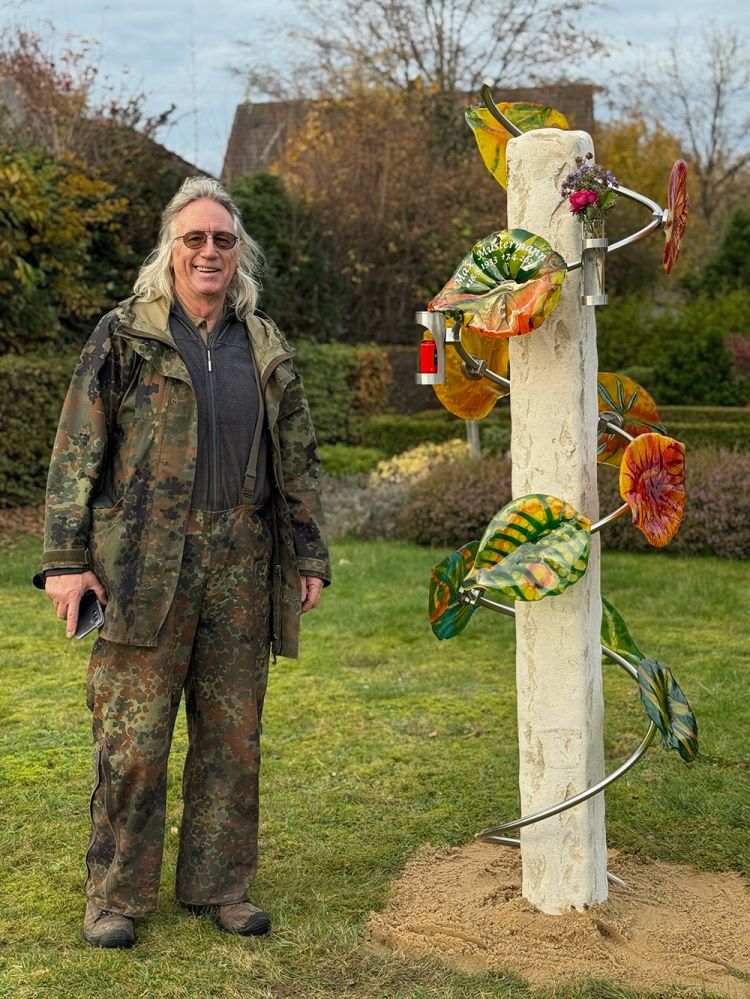 Person in Tarnkleidung steht neben einer weißen Säule mit bunten glasblättern und Blüten, die spiralförmig daran befestigt sind.