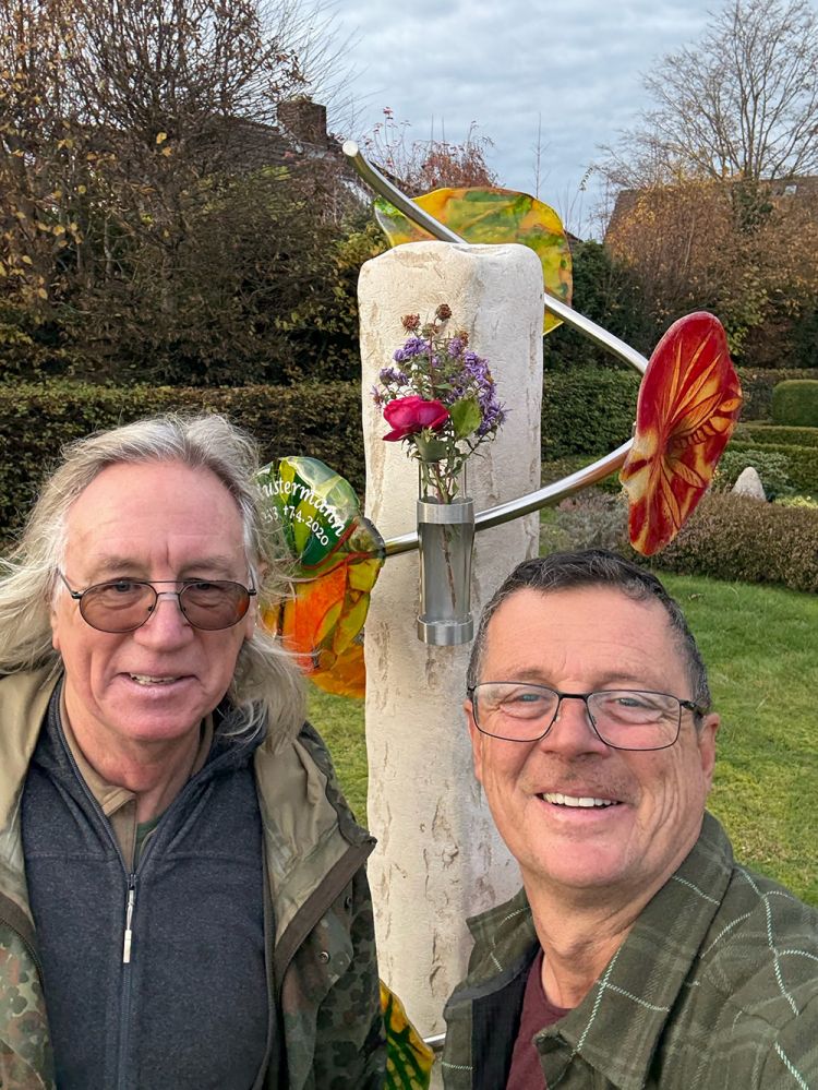 Steinerne Urnengemeinschaftsgrab-Säule mit Metallvase und buntem Glasornament auf den Friedhof, zwei Personen im Vordergrund.