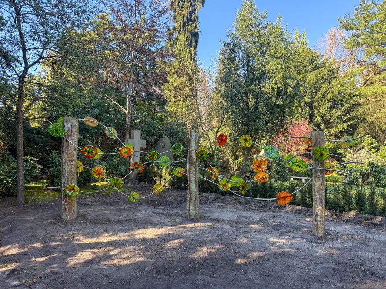 Urnen-Gemeinschafts-Grabanlage mit 40 bunten Glasblättern. Im Hintergrund ein Alter Grabstein und Bäume. Urnen-Gemeinschafts-Grabanlage mit 40 bunten Glasblättern. Im Hintergrund ein Alter Grabstein und Bäume.
